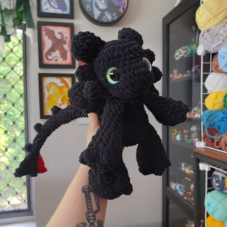 A black crocheted dragon being held up against a background of shelves and framed pictures. A black crocheted dragon being held up against a background of shelves and framed pictures.
