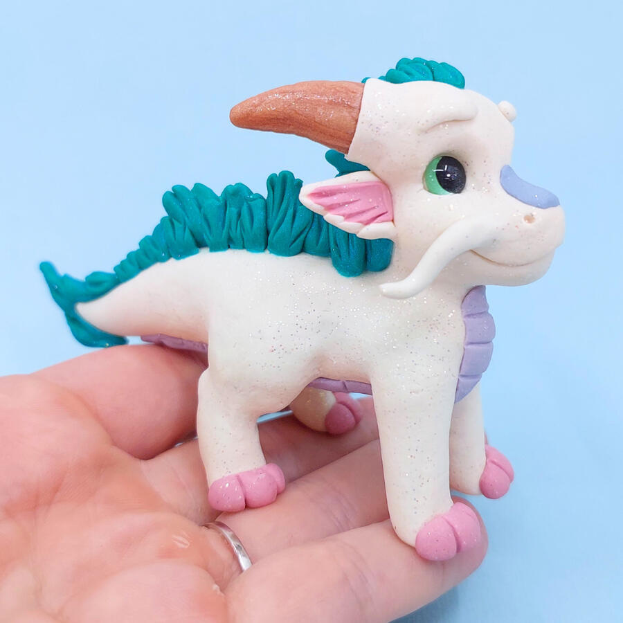 A white clay dragon standing on the fingers of an upturned hand in front of a light blue background. A white clay dragon standing on the fingers of an upturned hand in front of a light blue background.